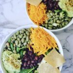 Two Green salads with guacamole, black beans, mixed greens, cheddar cheese, dried edamame and santa fe salad dressing in white bowls on a white countertop