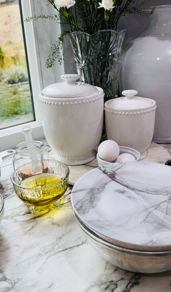 Walnut Roll Pastry with a glass bowl filled with yeast mixture fermenting with two white canisters a small glass bowl with oil and a bowl with eggs on a white countertop in front of a kitchen window.
