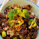 Asian Sweet Beef in a blue bowl chopsticks laying ontop of the rim of the bowl beef and broccoli with sesame seeds.