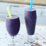 Blueberry Smoothie two cups filled with blueberry smoothie with a yellow and blue straw with spilled smoothie and chia seeds on a white countertop.