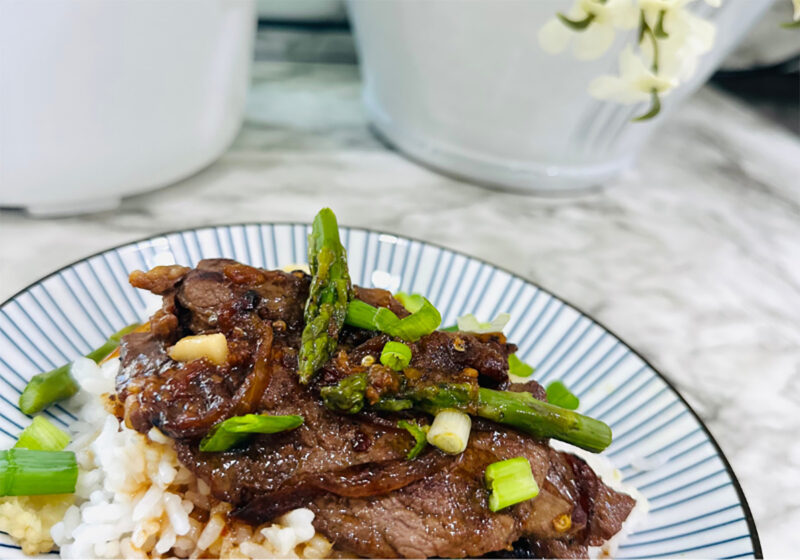 Mongolian Beef Asian on a white and blue striped platter with white vases and flower in background on white countops.