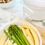 Steamed asparagus, baby corn, and bamboo shoots in a white plugin steamer with a bowl of onions on top of a wooden chopping board on a white countertop.