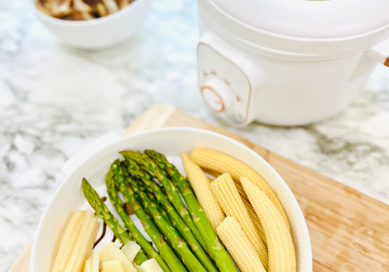Steamed asparagus, baby corn, and bamboo shoots in a white plugin steamer with a bowl of onions on top of a wooden chopping board on a white countertop.