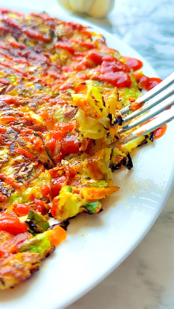 Our Flourless Healthy Cabbage Stir-fry pancakes! Cooked cabbage veggie pancake drizzled with siriracha sauce on a white plate and a white countertop with a garlic accenting photo top right of photo with fork.