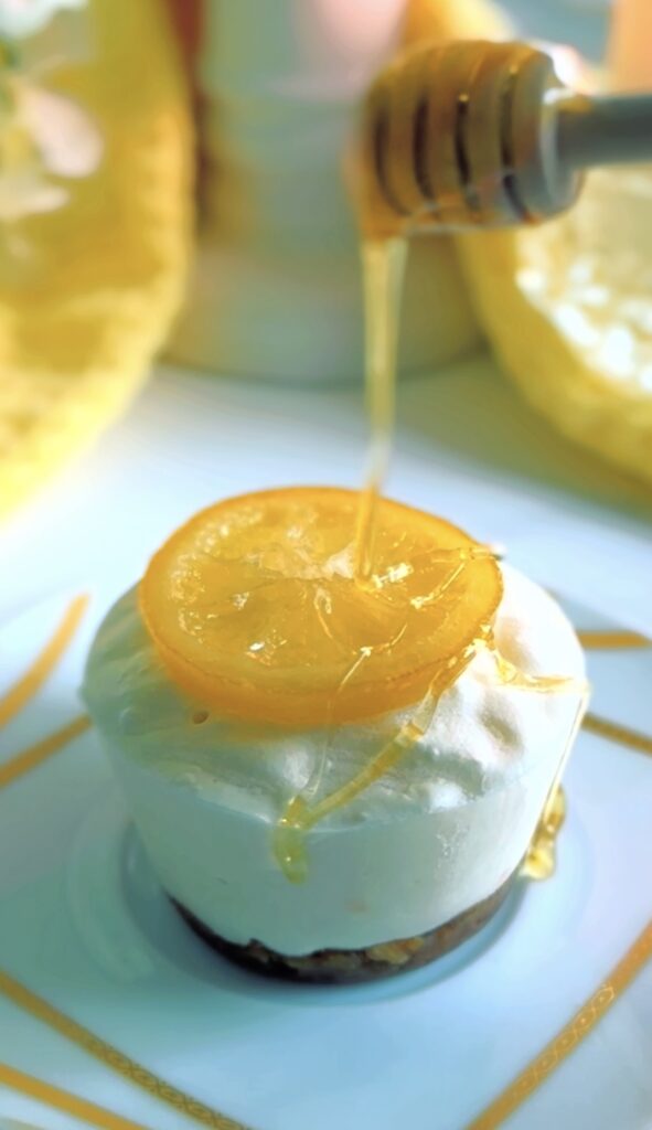 Lemon Cheesecake Cups on a white and gold sripped plate with honey drizzled on-top with cheesecakes lined up in the background on a white platter with a white dotted ceramic pitcher and a candle accented with some floral with a yellow towel.