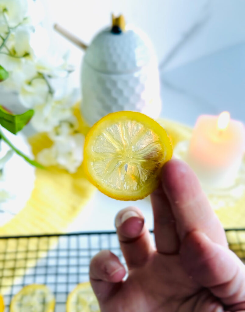 Candied Lemon Slice holding with a hand with a black cooling rack in the background with a lemon to the side of the bowl a white vase with flowers in the background with a candle lit up along with a honey jar in the background on a white countertop.