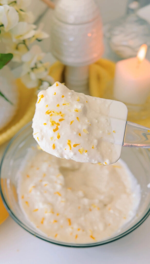 Glass bowl lemon zest over yogurt in a bowl with a lemon to the side of the bowl a white vase with flowers in the background with a candle lit up along with a honey jar in the background on a white countertop.