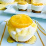 Lemon Cheesecake Cups on a white and gold sripped plate with honey drizzled on-top with cheesecakes lined up in the background on a white platter with a white dotted ceramic pitcher and a candle accented with some floral with a yellow towel.