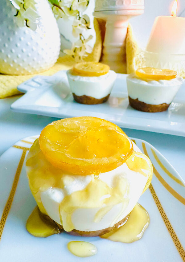 Lemon Cheesecake Cups on a white and gold sripped plate with honey drizzled on-top with cheesecakes lined up in the background on a white platter with a white dotted ceramic pitcher and a candle accented with some floral with a yellow towel.