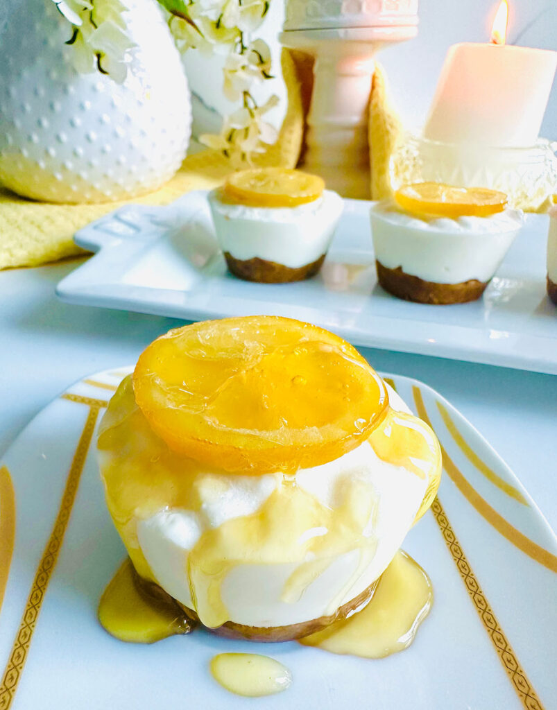 Lemon Cheesecake Cups on a white and gold sripped plate with honey drizzled on-top with cheesecakes lined up in the background on a white platter with a white dotted ceramic pitcher and a candle accented with some floral with a yellow towel.