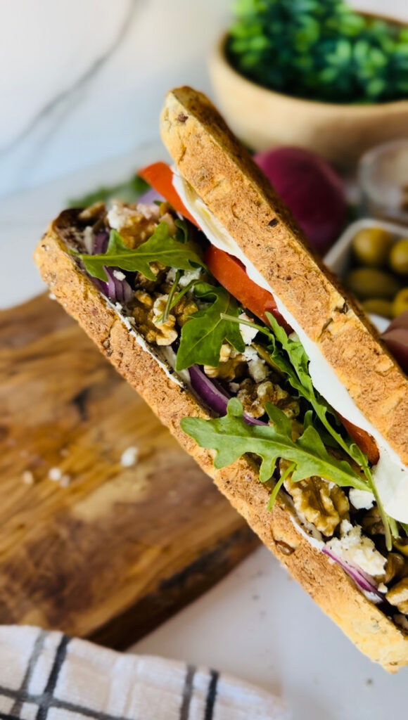 Rustic Greek Sandwhich - two slices of toasted bread added tomato slices, aurugla, walnuts, feta cheese ontop of a wooden chopping board accented with a grey stripped towel with a bowl of green mossy plant in back of sandwhich, in a white granet setting background.