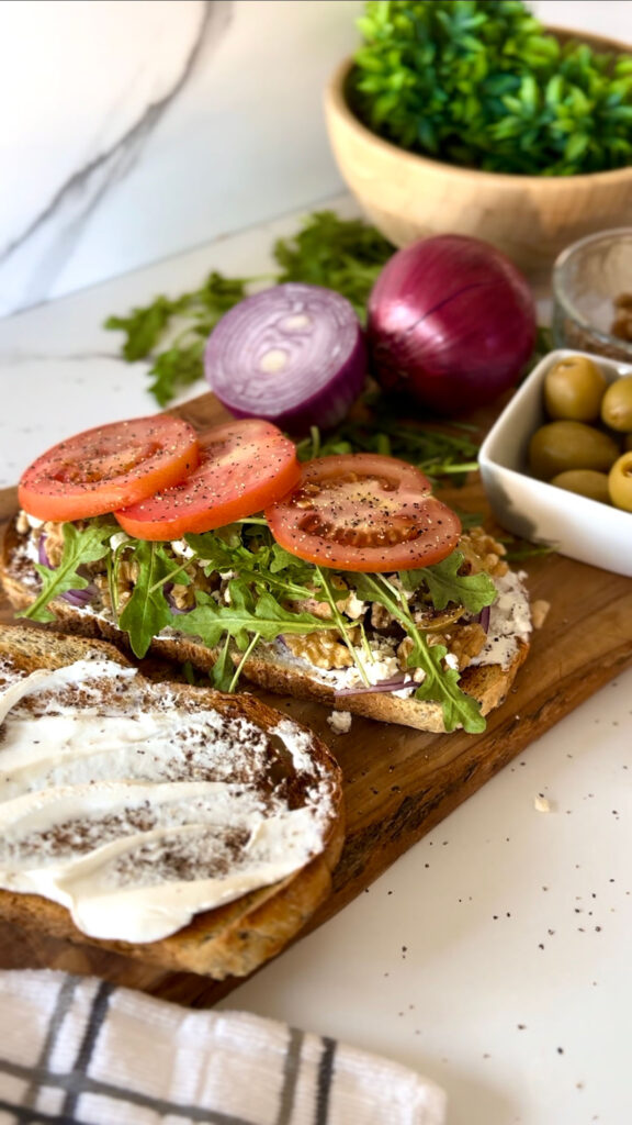 Rustic Greek Sandwhich - two slices of toasted bread added tomato slices, aurugla, walnuts, feta cheese ontop of a wooden chopping board accented with a grey stripped towel with a bowl of green mossy plant in back of sandwhich, in a white granet setting background.