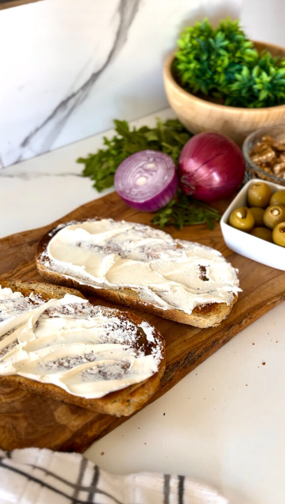Rustic Greek Sandwhich - two slices of toasted bread ontop of a wooden chopping board accented with a grey stripped towel with a bowl of green mossy plant in back of sandwhich, in a white granet setting background.