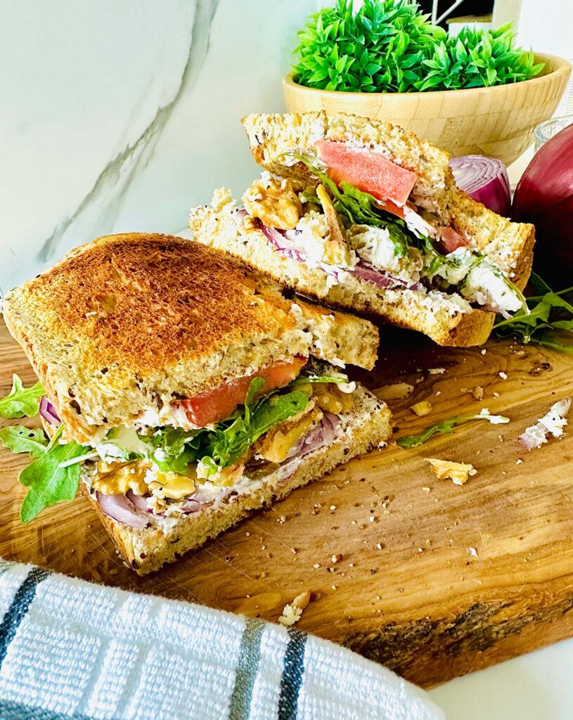Rustic Greek Sandwhich - cut sandwhich in half one layering the other ontop of a wooden chopping board accented with a grey stripped towel with a bowl of green mossy plant in back of sandwhich, in a white granet setting background.