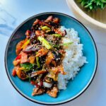 Blue round plate with soy glazzed eggplant garlic chicken stir-fry laying on a plate of white rice with a wooden bowl with green fern ball one a white countertop