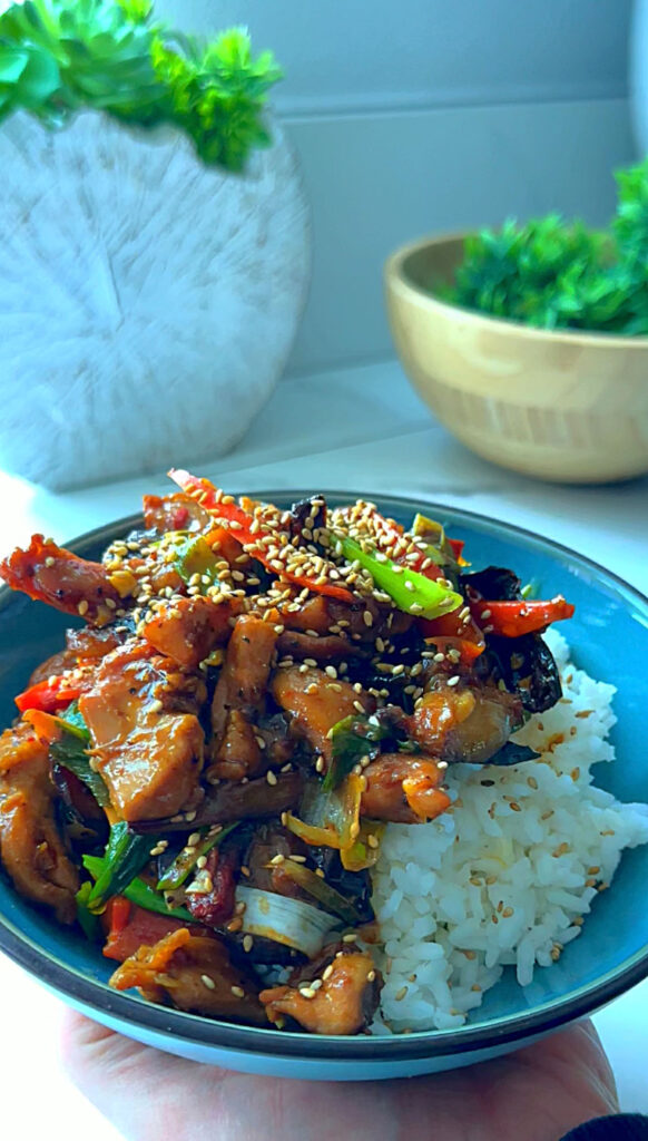 Blue round plate with soy glazzed eggplant garlic chicken stir-fry laying on a plate of white rice with a wooden bowl with green fern ball one a white countertop