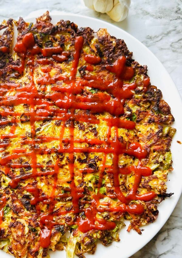 Our Flourless Healthy Cabbage Stir-fry pancakes! Cooked cabbage veggie pancake drizzled with siriracha sauce on a white plate and a white countertop with a garlic accenting photo top right of photo.