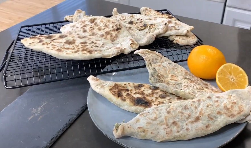 Zhingylov Hats- Flatbread stuffed with fresh green herbs and seasonings with a touch of lemon on a grey platter with fresh lemons accenting plate with a black cooling rack with flatbreads alighned to eachother. 