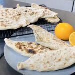 Zhingylov Hats- Flatbread stuffed with fresh green herbs and seasonings with a touch of lemon on a grey platter with fresh lemons accenting plate with a black cooling rack with flatbreads alighned to eachother.