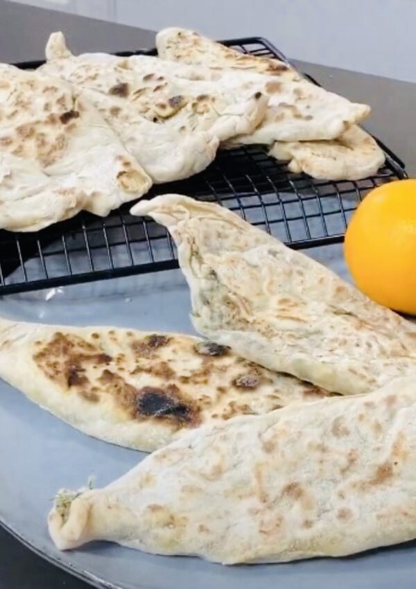 Zhingylov Hats- Flatbread stuffed with fresh green herbs and seasonings with a touch of lemon on a grey platter with fresh lemons accenting plate with a black cooling rack with flatbreads alighned to eachother.