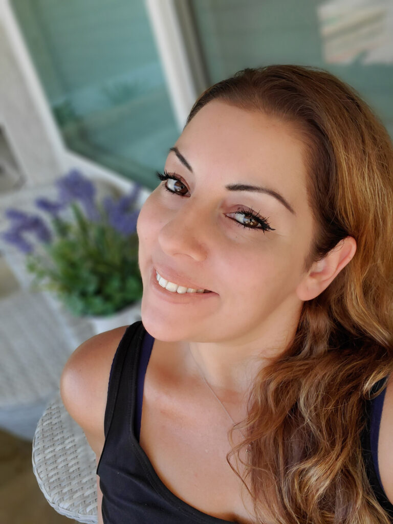 Galore Of Flavors Sylvia Creator with a black shirt with purple lilac flowers in a white pitcher outdoor setting with windows behind her.