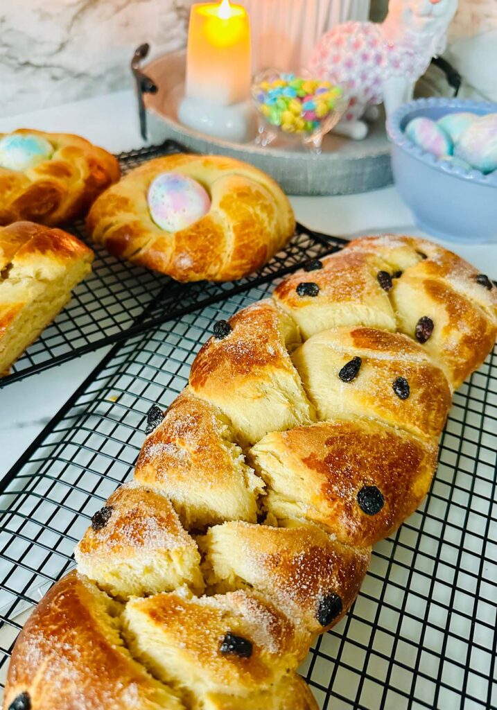 Sweet easter bread with dried cranberries on a black colling rack with two round shaped breads accented with a colored easter egg in the center with a blue bowl of tie-dye pastel colored easter eggs with a gray tray with a lit candle in the background.