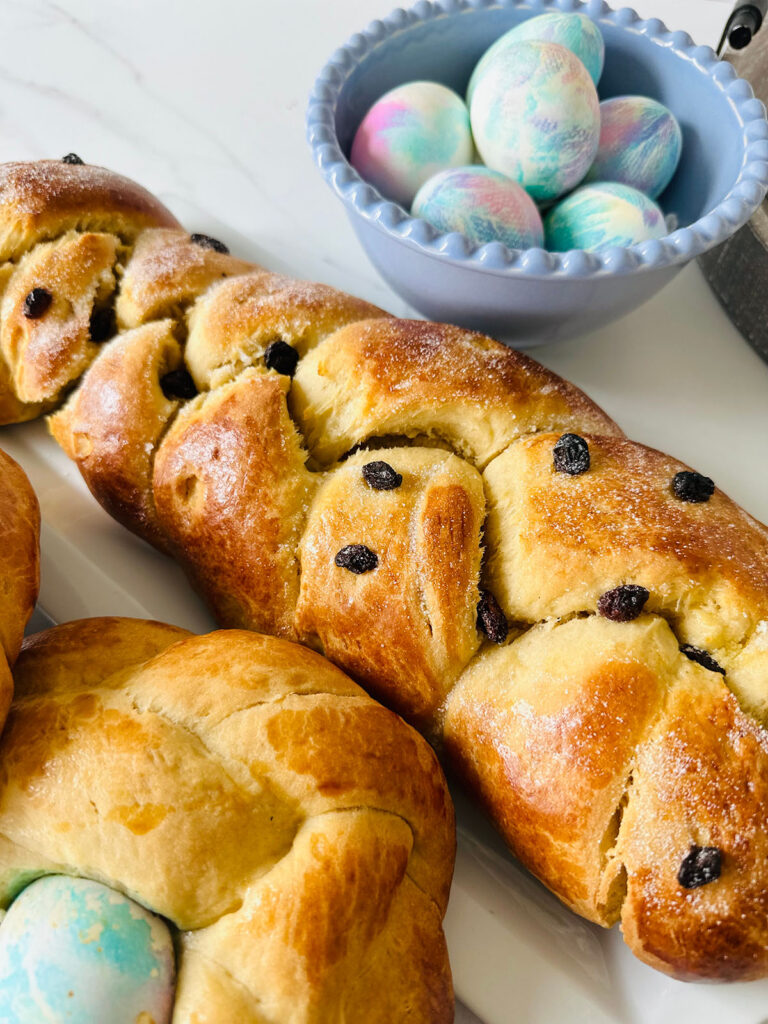 Sweet easter bread with dried cranberries on a black colling rack with two round shaped breads accented with a colored easter egg in the center with a blue bowl of tie-dye pastel colored easter eggs with a gray tray with a lit candle in the background.
