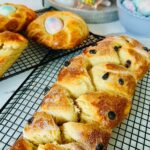 Sweet easter bread with dried cranberries on a black colling rack with two round shaped breads accented with a colored easter egg in the center with a blue bowl of tie-dye pastel colored easter eggs with a gray tray with a lit candle in the background.