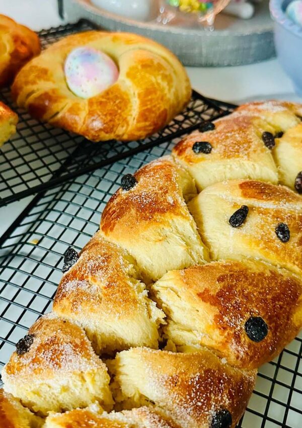 Sweet easter bread with dried cranberries on a black colling rack with two round shaped breads accented with a colored easter egg in the center with a blue bowl of tie-dye pastel colored easter eggs with a gray tray with a lit candle in the background.