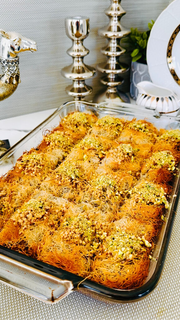 Kadaif Kunafa dessert in a glass baking tray with crushed walnuts and pastacios with a silver accented decor of a camel, silver candle sticks and a white with black and gold patterned pitcher pourer with a white plate and green plant in a grey vase in background.