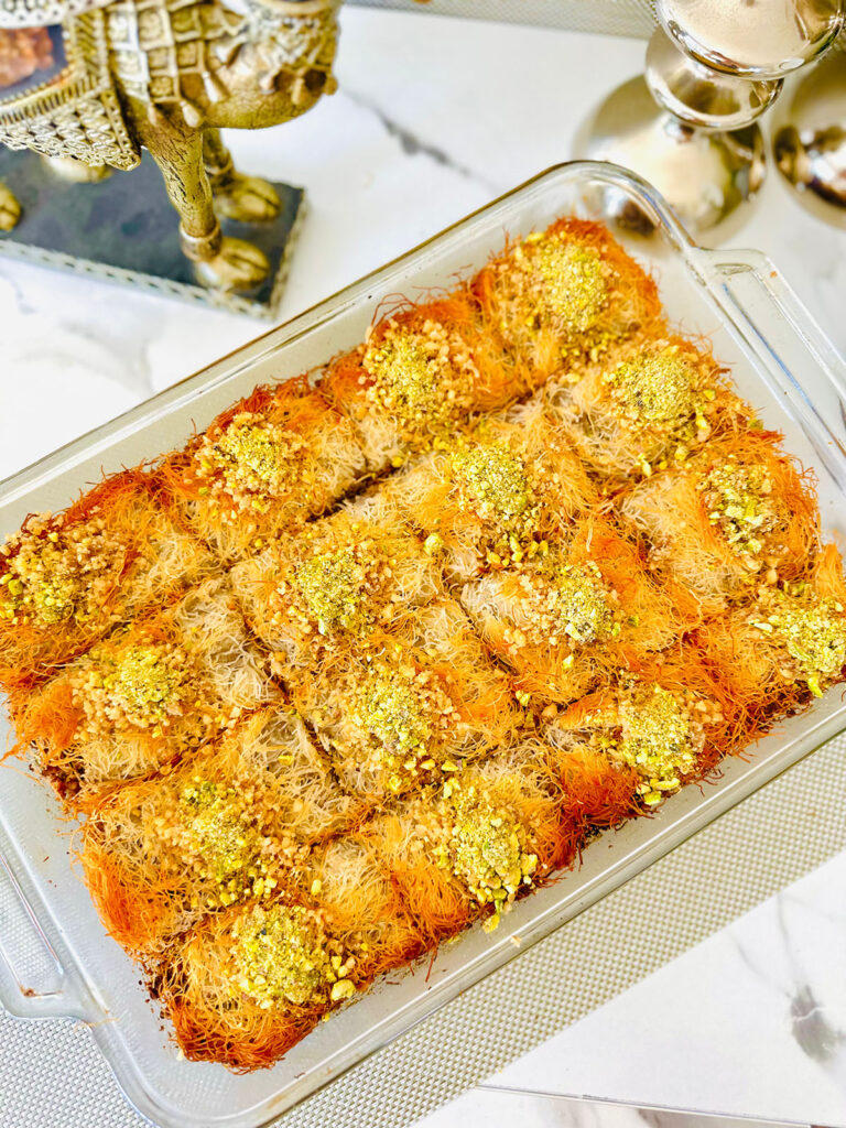 Kadaif Kunafa dessert in a glass baking tray with crushed walnuts and pastacios with a silver accented decor of a camel, silver candle sticks and a white with black and gold patterned pitcher pourer with a white plate and green plant in a grey vase in background.