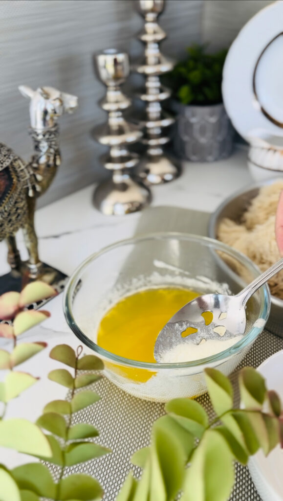 Skimming white film from butter clarifying butter in a glass round bowl on wooden chopping board with accent decor or a silver camel and 2 candle sticks with a grey plant pot in background with a white gold and black pitcher on a white marble countertop.