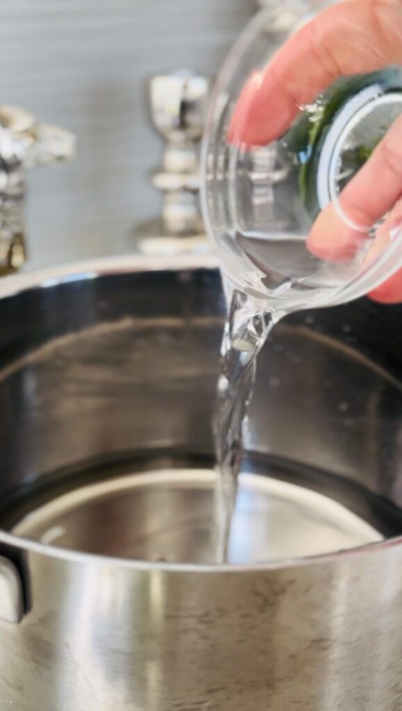 stainless steal pot filled with water pouring rose water with a small glass bowl with accent decor or a silver camel and 2 candle sticks with a grey plant pot in background with a white gold and black pitcher on a white marble countertop.