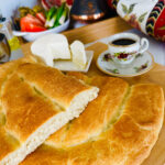 Flatbread cut in half on a wooden chopping board with feta cheese in a white plate next to a cup of coffee. Platter of vegetables in the background.