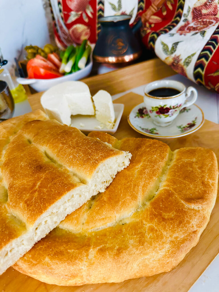 Flatbread cut in half on a wooden chopping board with feta cheese in a white plate next to a cup of coffee. Platter of vegetables in the background.