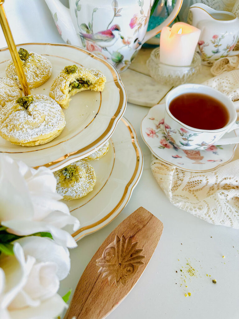 Ma'amoul Pistachio Cookie on a two tear cookie white platter with gold trimming a ma'amoul cookie wooden mold maker cream roses accenting photo on left side with a floral tea cup china set with a matching china tea pot in background with a lit candle with a crystal candleholder and lace cloth accenting table on a white countertop