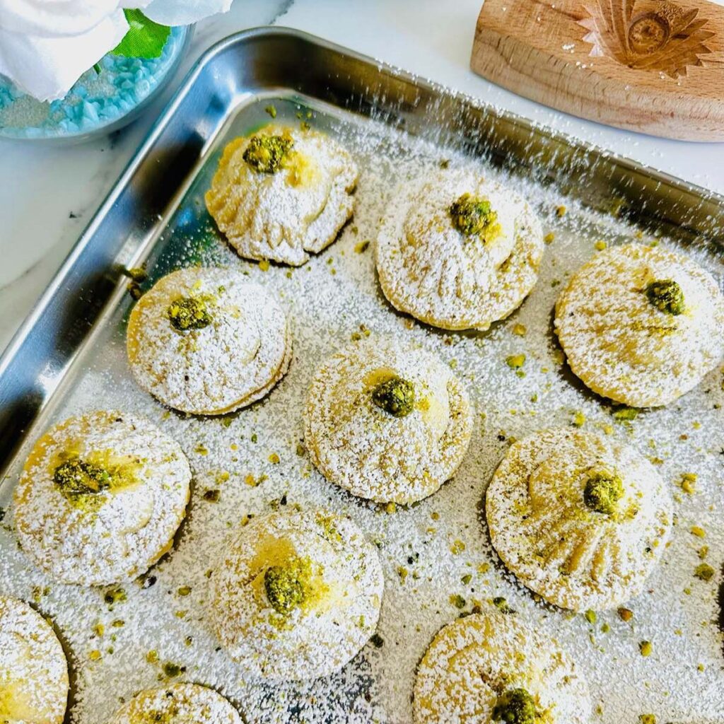Ma'amoul Pistachio Cookie on a silver tin baking sheet with white powdered sugar sprinkled over ma'amoul cookies lined up with a vase of cream colored flowers in photo