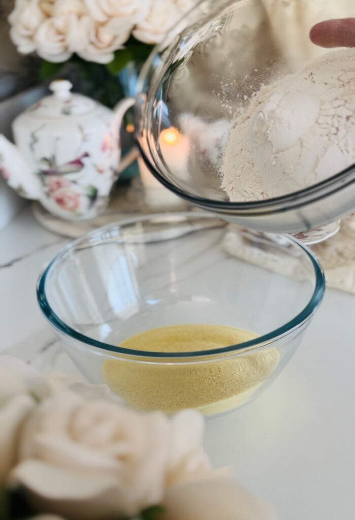 Two glass bowls one filled with semolina and the other filled with flour pouring into bowl with semolina cream roses accenting photo on left side with a floral tea cup china set with a matching china tea pot in background with a lit candle with a crystal candleholder and lace cloth accenting table on a white countertop
