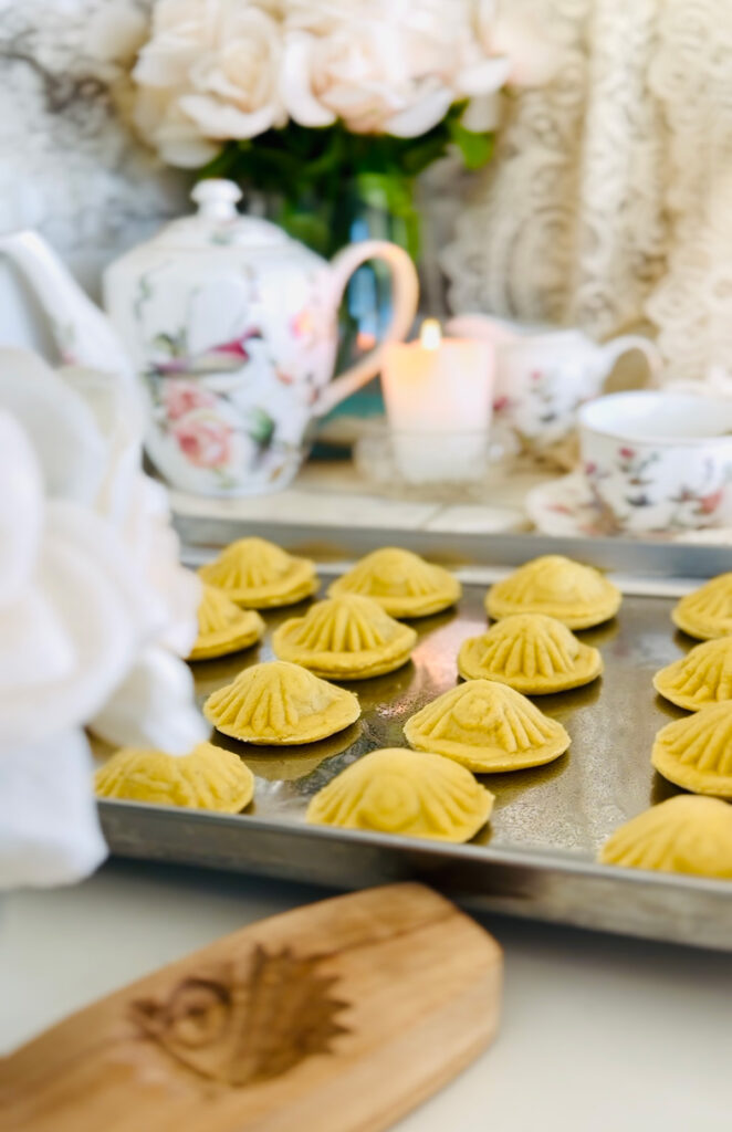 Ma'amoul Pistachio Cookie on a silver tin baking sheet cookie shapped with mold and lined 1" apart from eachother ma'amoul cookies lined up with a vase of cream colored flowers in photo with a tea set in background with floral and birds blurred in the background with a candle light on a white countertop