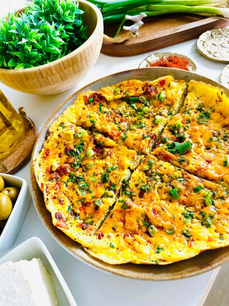 Tomato Egg Scramble on a round platter with olive and feta cheese in a white bowl, green plant in a wooden bowl with a wooden tray with two golden birds in background with a bottle of oil and red pepper in golden seasoning plate