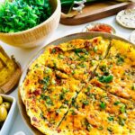 Tomato Egg Scramble on a round platter with olive and feta cheese in a white bowl, green plant in a wooden bowl with a wooden tray with two golden birds in background with a bottle of oil and red pepper in golden seasoning plate
