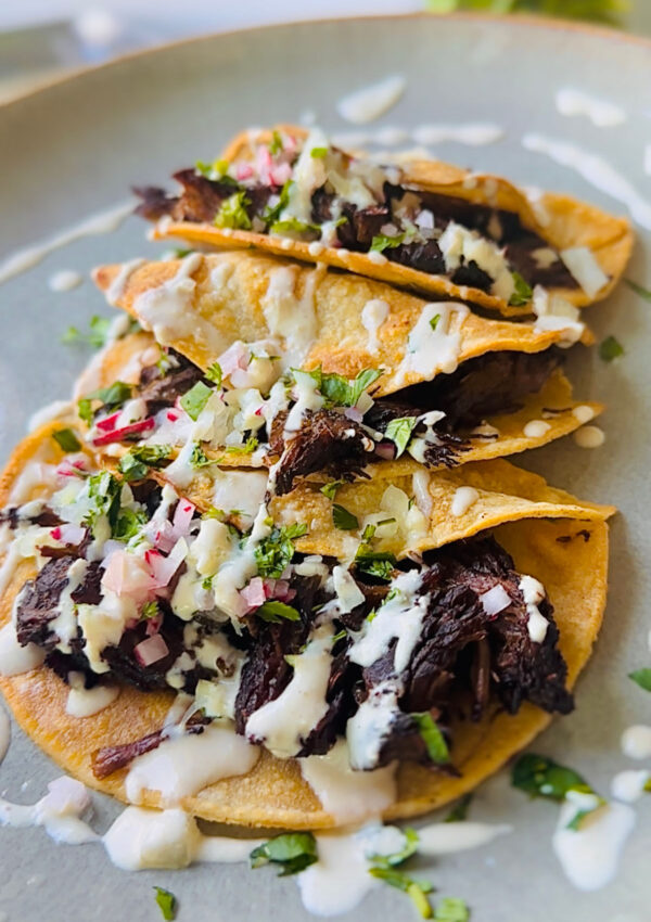 Beef Shawarma Tacos - 3 tacos laying in a plate topped with chopped raddish, onions and cilantro with tahini sauce drizzled over with a green plant in backbround on a white countertop.