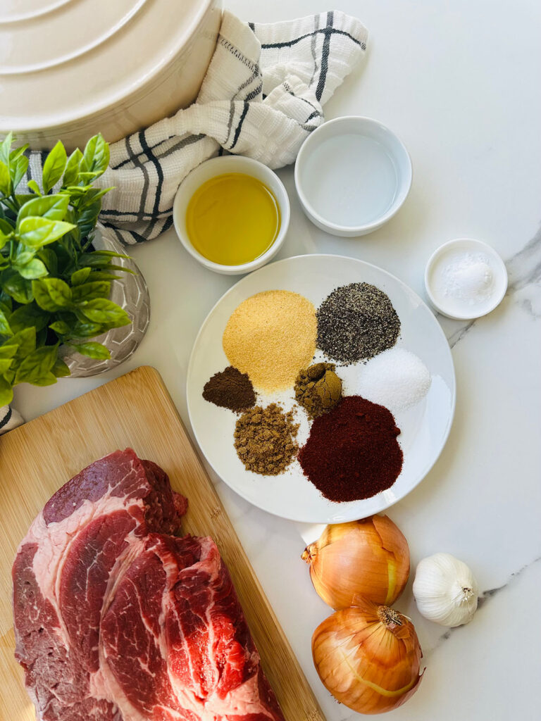 Beef Shawarma Tacos - beef on a wooden chopping board, seasonings on a white plate onions and garlic with white bowls filled with oil, vinegar and salt with a green land in a grey pottery with a lined towel and a stone baking pot on a white countertop