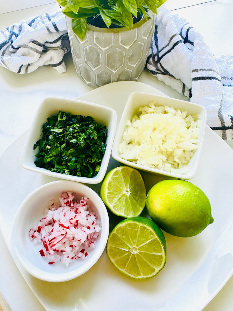 Beef Shawarma Tacos - White platter with white plates of chopped cilantro, onions, raddish cut limes in half with a green land in a grey pottery with a lined towel and a stone baking pot on a white countertop
