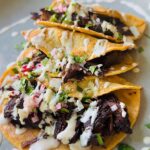 Beef Shawarma Tacos - 3 tacos laying in a plate topped with chopped raddish, onions and cilantro with tahini sauce drizzled over with a green plant in backbround on a white countertop.