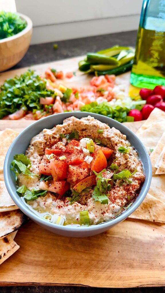 Foul (Ful) Medamas - Fava Bean Stew Ful in a blue bowl topped with diced tomatoes, cilantro and green onions accented on top. Chopped vegetables in the background with cut up pita bread surrounding the blue bowl on a wooden chopping board.