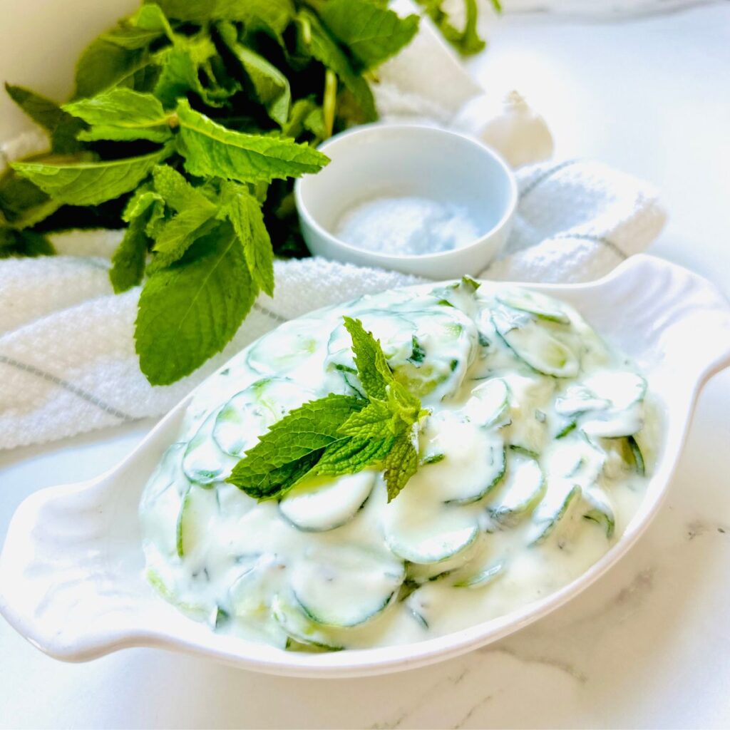 Jajukh | Refreshing Yogurt Salad with Cucumbers and Mint in a white long meza plate with yogurt cucumbers inside plate accented with a fresh mint leave with a pile of cucumbers in backbround laying ontop of a white stripped kitchen towel.