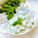 Jajukh | Refreshing Yogurt Salad with Cucumbers and Mint in a white long meza plate with yogurt cucumbers inside plate accented with a fresh mint leave with a pile of cucumbers in backbround laying ontop of a white stripped kitchen towel.