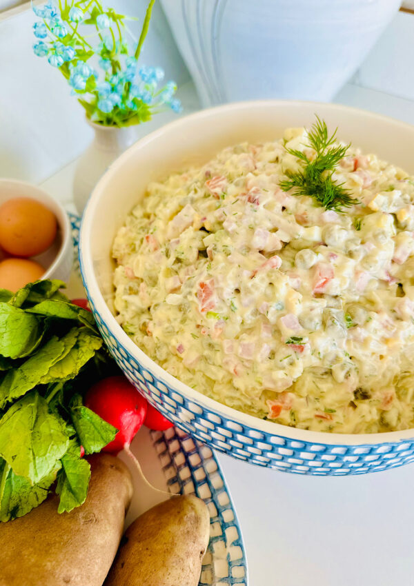 Russian Olivier Salad - Simple Potato Salad in a blue patterend bowl accented with fresh dill with raddish with green leaves and brown eggs in a platter on the side with a blue accented plant in background.