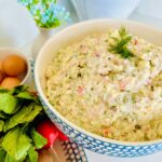 Russian Olivier Salad - Simple Potato Salad in a blue patterend bowl accented with fresh dill with raddish with green leaves and brown eggs in a platter on the side with a blue accented plant in background.